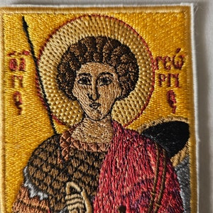以下が含まれることがあります： 聖人が槍を持っている刺繍パッチ。黄色い背景に聖人の頭上に後光が差しています。パッチには「S.T.G」と「REG.P.H.」の文字が見えます。