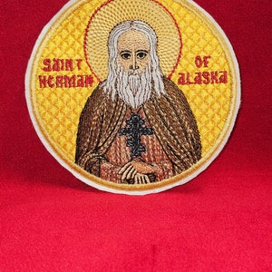 May include: Embroidered patch featuring a yellow background with a saint in a brown robe. The saint has a white beard and is holding a cross. The text "SAINT HERMAN OF ALASKA" is embroidered around the saint.