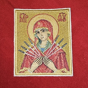 Puede incluir: Parche bordado de icono religioso que representa a una mujer con un halo, con las manos juntas, con siete espadas que le atraviesan el pecho. El icono está enmarcado en oro con texto en las esquinas superiores, sobre un fondo rojo.