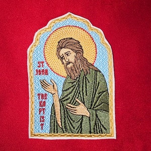 May include: Embroidered patch depicting a religious icon of Saint John the Baptist. The patch is in a traditional style with a blue and gold background and a halo around the saint's head. The text "ST JOHN THE BAPTIST" is embroidered on the left side of the patch.