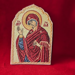 May include: Embroidered icon of a woman in a red robe with a gold halo. The icon is on a white background with a gold border.