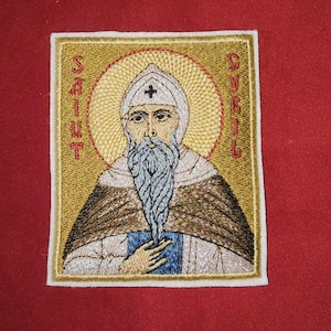 May include: Embroidered patch featuring Saint Cyril, with a gold halo and the text "SAINT CYRIL" in red. The saint is depicted with a white beard, a white hat with a cross, and a brown cloak. The patch is bordered with a white frame.