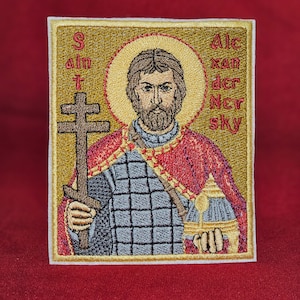 Op de afbeelding: Gestikte patch met een religieus icoon van een man met een baard die een kruis vasthoudt. De achtergrond is goud met een rode en witte halo rond het hoofd van de man. De tekst "Sint Alexander Nevski" is in rood gestikt aan de rechterkant van de patch.