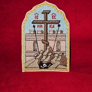 Könnte beinhalten: Ein gestickter Aufnäher mit blauem Hintergrund und goldener Umrandung. Der Aufnäher zeigt ein braunes Kreuz mit den kyrillischen Buchstaben "IC XC" darüber und "НИ КА" darunter. Das Kreuz steht vor einem weißen Gebäude mit einem Totenkopf und gekreuzten Knochen an der Basis.