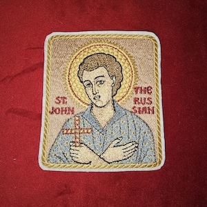Może przedstawiać: Naszywka haftowana przedstawiająca świętego z aureolą, trzymającego krzyż. Naszywka jest beżowa z złotą obwódką i tekstem "ST JOHN THE RUSSIAN" w kolorze czerwonym.