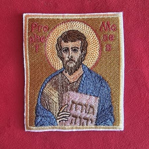 Könnte beinhalten: Gestickter Aufnäher mit einer religiösen Figur mit Heiligenschein, die ein Buch hält. Die Figur hat einen Bart und trägt blaue und braune Gewänder. Der Hintergrund ist goldfarben mit Text oben. Der Aufnäher ist quadratisch mit weißem Rand.