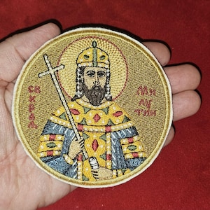 May include: Embroidered patch featuring a religious figure with a halo, holding a cross. The patch has gold, blue, and yellow colors, with text in a foreign language. The patch is round and has a cream-colored border.