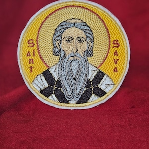 Pode incluir: Etiqueta bordada com um design circular amarelo e branco com uma representação de São Sava. A etiqueta tem as palavras "Saint" e "Sava" escritas verticalmente nos lados esquerdo e direito da imagem.