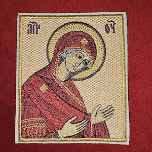 May include: Embroidered patch featuring a religious icon of a woman with a halo, wearing a red robe and head covering. The image has gold accents and text in the upper corners. The patch is square with a white border.