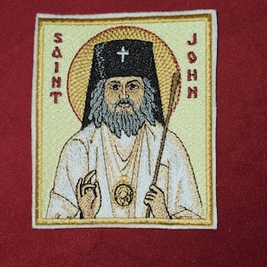 Może przedstawiać: Haftowana naszywka przedstawiająca ikonę Świętego Jana. Obraz przedstawia brodatego mężczyznę w czarnym kapeluszu z krzyżem, trzymającego laskę. Napis "SAINT JOHN" jest wyhaftowany złotą nicią wokół obrazu na czerwonym tle.