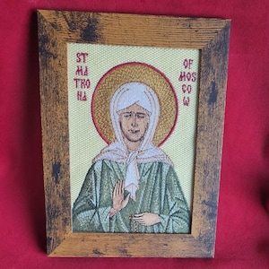 May include: Embroidered religious icon of a woman in a green robe and white head covering, framed in a rustic wooden frame. The icon features text in red and a gold halo. The background is a light yellow color.