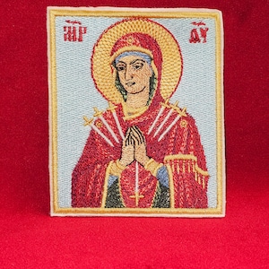 May include: Embroidered patch featuring a woman in a red robe with a halo, holding her hands in prayer. The woman is surrounded by seven swords. The patch has a gold frame and the text "MP" and "OY" in the corners.