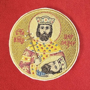 May include: Embroidered patch featuring a religious figure with a halo, crown, and cross. The figure is depicted in gold, red, and blue hues, holding a smaller figure. Text in Cyrillic script is visible.