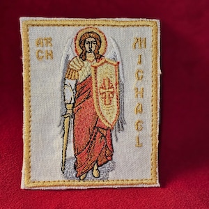 Op de afbeelding: Gestikte patch met een witte achtergrond en een gouden rand. De patch toont een engel met een zwaard en schild, met de tekst "ARCH MICHAEL" in goud geborduurd aan de rechterkant.