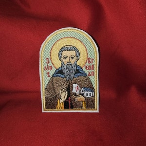 Op de afbeelding: Gestikte patch met een heilige in een bruine robe die een klein gebouw vasthoudt. De heilige heeft een grijze baard en een halo. De patch heeft een gouden rand en de tekst "St.  " en "ET" aan weerszijden van de heilige.