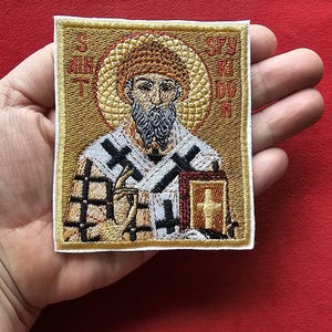 May include: Embroidered patch featuring a religious figure with a halo, beard, and cross-adorned book. The patch has a gold background with detailed stitching in various colors. The text "S. SPYRIDON" is visible around the halo. The patch is square with a white border.