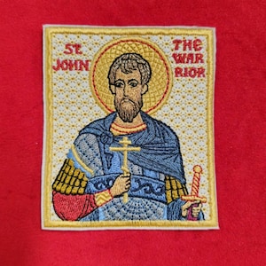 May include: Embroidered patch depicting Saint John the Warrior. The patch is beige with a gold border and features a white background with a gold pattern. The image shows Saint John holding a sword and a cross. The text "St John" is in the top left corner and "The Warrior" is in the top right corner.