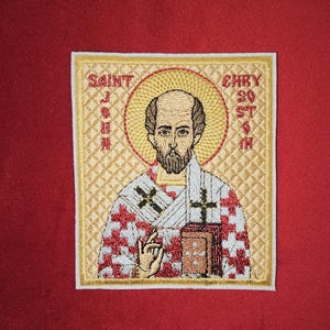 May include: Embroidered patch featuring a saint with a red and white checkered robe, holding a book. The patch has a gold border and the text "SAINT JOHN CHRYSOSTOM" is embroidered around the image.