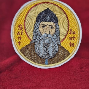 May include: Embroidered patch featuring a saint with a beard and a black hat. The patch is yellow with a white border and the words "Saint Justin" are embroidered on the side.