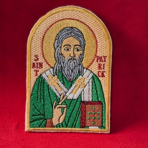 Op de afbeelding: Geborduurde patch met Sint-Patrick, afgebeeld met een aureool, groene gewaden en een boek. De patch heeft de tekst "SAINT PATRICK" en is geplaatst tegen een rode achtergrond.
