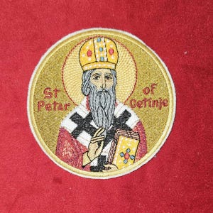 Könnte beinhalten: Gestickter Aufnäher mit einem religiösen Bildnis des Heiligen Petar von Cetinje. Die Ikone zeigt einen bärtigen Mann mit Krone und rotem Gewand, der ein Buch hält. Der Aufnäher ist rund und hat einen goldenen Hintergrund mit einem weißen Rand.
