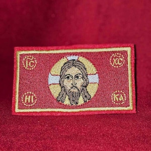 May include: A red embroidered patch with a gold border. The patch features a stylised image of Jesus Christ with a halo and a beard. The letters "IC XC" are embroidered in gold in the corners of the patch.