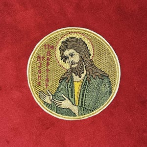 Op de afbeelding: Ronde geborduurde patch met Johannes de Doper met een gouden aureool. De patch heeft gedetailleerd borduurwerk in groen, geel en bruin. De tekst "The St. John" is zichtbaar.