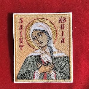 May include: Embroidered patch featuring a religious icon of a saint. The image depicts a woman with a halo, wearing a green robe and white head covering. The words "SAINT" and "XSHIA" are embroidered in red.