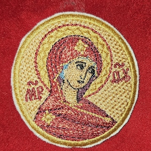 May include: A circular embroidered patch featuring a religious icon of a figure in a red head covering and robe, surrounded by a gold halo. The design includes red Greek lettering on both sides and a white border, set against a solid red background.