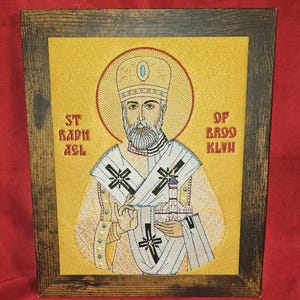 May include: Embroidered icon of St. Raphael of Brooklyn in a wooden frame. The image features a man with a beard, wearing a religious garment with crosses. The background is yellow with text that reads "ST RAPHAEL OF BROOKLYN".