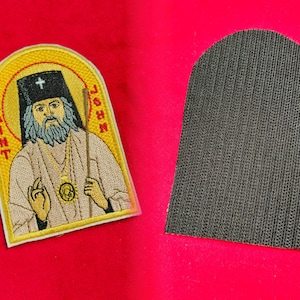 Może przedstawiać: Naszywka haftowana do prasowania z żółtą ramką z napisem "Saint John" i obrazem brodatego mężczyzny w białej szacie i złotym krzyżu.