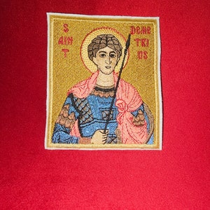 Op de afbeelding: Gestikte patch met een heilige die een speer vasthoudt, met de tekst "SAINT" en "DEAC TRIS" aan de linker- en rechterkant. De patch is op een rode achtergrond.