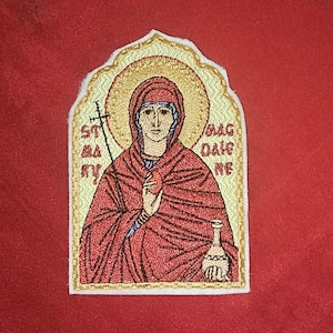 Op de afbeelding: Geborduurde patch met een religieus icoon van een vrouw in een rode jurk, die een hart en een fles vasthoudt. De afbeelding heeft een gouden halo en is omlijst door een gouden rand. De tekst in de afbeelding luidt "ST MARY MAGDALENE".