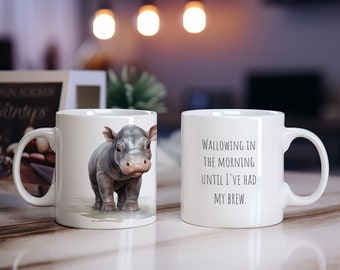 May include: Two white ceramic mugs. One mug features a cartoon illustration of a baby hippopotamus. The other mug has the text "Wallowing in the morning until I've had my brew."
