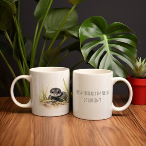 May include: Two white ceramic mugs, one with a snake illustration and the other with the text "Hiss-terically in need of caffeine!"