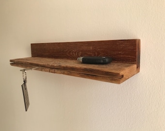 Può includere: Un ripiano in legno rustico con un gancio per le chiavi e un piccolo oggetto nero in cima. Il ripiano è realizzato in legno riciclato e ha una finitura naturale.