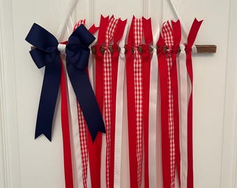 May include: A patriotic wall hanging featuring red, white, and blue ribbons. The ribbons are attached to a wooden dowel and adorned with a navy blue bow. Some ribbons have a gingham pattern.