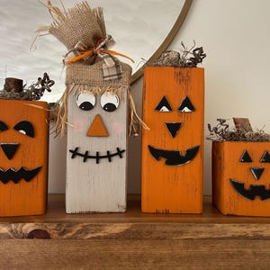 May include: Four wooden Halloween decorations. Two orange jack-o'-lanterns, a scarecrow, and a small pumpkin. The jack-o'-lanterns have black triangle eyes and carved smiles. The scarecrow has a burlap hat and straw hair. The decorations are on a wooden shelf.
