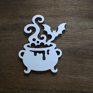 May include: A light-colored wooden cutout featuring a cauldron with liquid bubbling over the rim, accompanied by swirling steam and a small bat silhouette rising from the top. The design is flat with smooth edges and a natural wood grain texture.