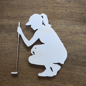 May include: A white wooden cutout silhouette of a golfer in a cap, crouching down while holding a golf putter. The figure is depicted in a side profile view, showing the golfer leaning forward to inspect the ground with the putter held vertically.