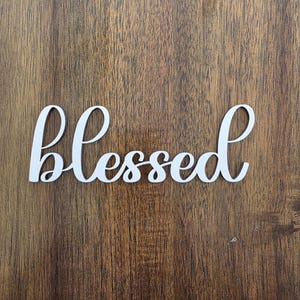 May include: White cursive word "Blessed" on a wooden background. The word is in a flowing script style, with the letters connected. The wood has a dark brown color with visible grain.
