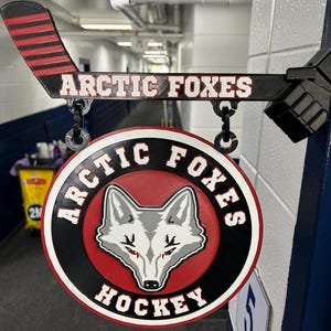 Peut inclure: Une enseigne murale noire et rouge en forme de crosse de hockey avec le texte ARCTIC FOXES. Un panneau circulaire est suspendu en dessous, affichant un graphique de tête de renard stylisé au centre avec le texte ARCTIC FOXES HOCKEY sur le bord.