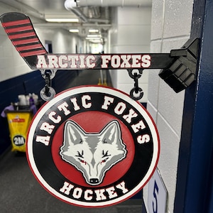 May include: A wall-mounted sign featuring a black hockey stick with red stripes and the text ARCTIC FOXES. A circular sign hangs from the stick, displaying a grey fox head graphic, a red background, and the text ARCTIC FOXES HOCKEY.