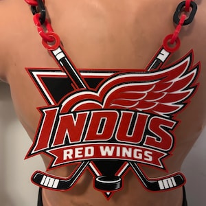 以下が含まれることがあります： 「INDUS RED WINGS」という文字と交差したホッケーのスティック、赤い翼のあるホイールのロゴが描かれた、大きく平らな装飾用ペンダントネックレス。ペンダントは、赤と黒のプラスチック製のリンクが交互に連なった太いチェーンから吊り下げられています。