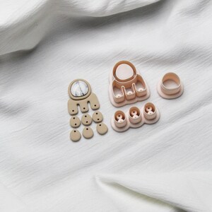 May include: A set of polymer clay earring making supplies. The set includes a variety of shapes and sizes, including a large round shape, a smaller round shape, and several smaller oval shapes. The shapes are all in a light pink color.