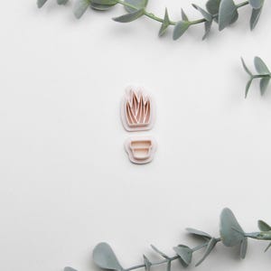 May include: A set of two white plastic cookie cutters shaped like a potted plant. The plant cutter has a simple design with five leaves. The pot cutter has a rounded shape with a single line around the top.
