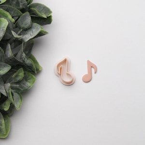 May include: Two light pink musical note-shaped cookie cutters. One is a single note, and the other is a double-layered cutter. The cutters are on a white surface with green foliage in the upper left corner.