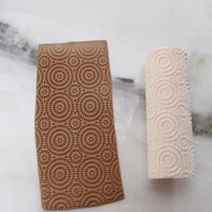 May include: Two polymer clay tools with circular patterns. One is a rectangular brown clay slab with a repeating circle design. The other is a cylindrical off-white clay roller with a spiral pattern. Both tools are for creating textured surfaces.