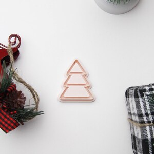 May include: A light pink, layered Christmas tree-shaped cookie cutter. The cutter is surrounded by festive decorations, including a miniature Christmas tree, a red sled with pine and a plaid wrapped gift.