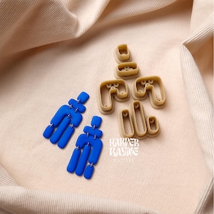 May include: Two pairs of blue polymer clay earrings with a geometric design. The earrings are displayed on a beige fabric surface with the moulds used to create them.